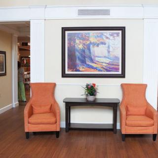 A cozy living room area featuring two orange chairs, a dark wooden table with a small potted plant, and a colorful framed painting on the wall.