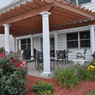 A backyard patio area featuring a wooden pergola supported by white columns, surrounded by lush landscaping and flowering plants.
