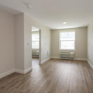 A bright, empty apartment room with windows and wooden flooring.