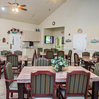 A cozy dining room with multiple tables and chairs, decorated with plants and featuring a small kitchen area and a television.