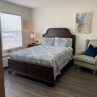 A cozy bedroom with a neatly made bed, a lamp on the bedside table, an armchair, and artwork on the wall.