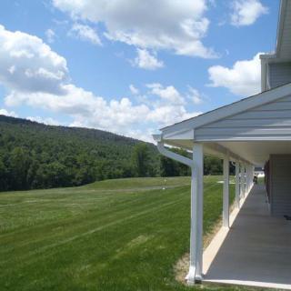 A serene view of a house with a porch overlooking lush green fields and distant hills under a partly cloudy sky.