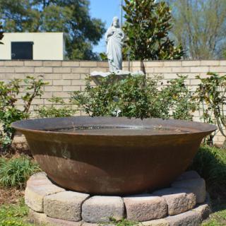 A large, round fountain in a garden setting with a statue and brick wall in the background.