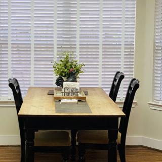 A cozy dining area with a wooden table and chairs, decorated with a flower arrangement near a window with blinds.