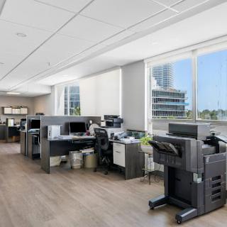 A modern office space featuring cubicles with computers and printers, illuminated by natural light coming through large windows.