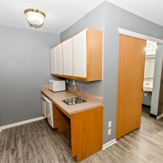 A small kitchen area with wooden cabinets and a sink, featuring a microwave on the countertop and a doorway leading to another room.