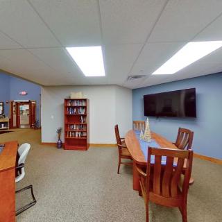 A modern conference room with a long table, chairs, a large television, and a bookshelf.