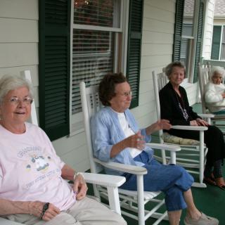 A group of elderly women sitting and conversing on a porch in rocking chairs.