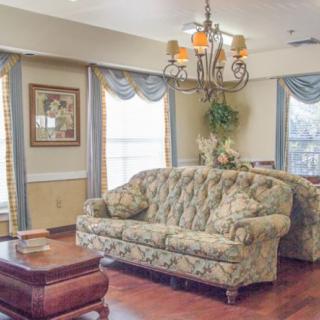 A cozy living room with a floral sofa, elegant chandelier, and large windows adorned with curtains.