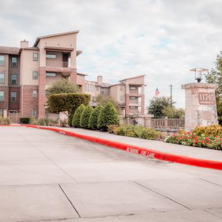 A view of a residential apartment complex with a driveway and landscaped surroundings under a cloudy sky.
