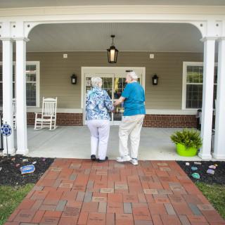 Two elderly individuals walking together towards a home's entrance, showcasing companionship and support.
