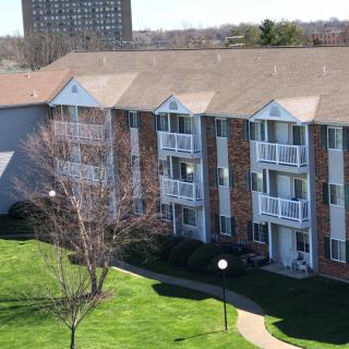 A multi-story apartment building with brick exterior and balconies, surrounded by green lawns and trees.