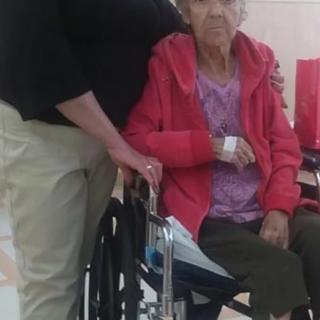 A caregiver stands next to an elderly woman sitting in a wheelchair, likely in a hospital setting.
