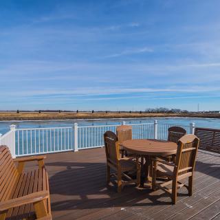 A scenic view of a waterfront deck with wooden chairs and table under a clear blue sky.