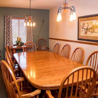 A cozy dining room with a large wooden table and matching chairs, illuminated by warm lighting fixtures and complemented by wall art and curtains.