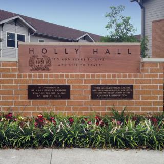 A sign on a brick wall for Holly Hall Retirement Community, featuring plants in front.