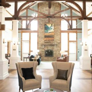 A cozy living room with two armchairs facing a stone fireplace, flanked by large windows and exposed wooden beams.