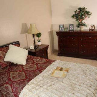 A cozy bedroom featuring a bed with patterned bedding, a nightstand with lamps, and a dresser adorned with photos and decor.