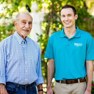 Two men smiling outdoors, one in a checkered shirt and the other in a teal polo.