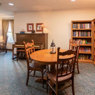 A cozy living room featuring a wooden dining table, chairs, a bookshelf filled with books, and a piano.