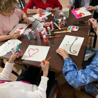 A group of people engaging in a painting activity, creating heart and swirl designs on paper at a table.