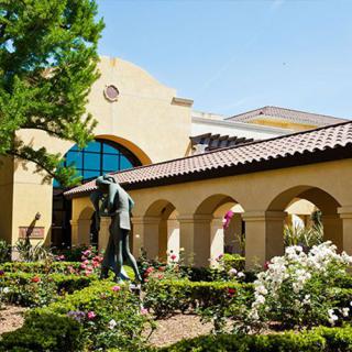 A beautiful building with arches, surrounded by a lush garden featuring a statue and blooming flowers.