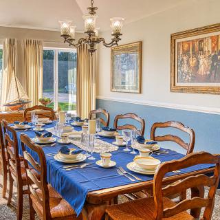 A well-appointed dining room featuring a wooden table set with a blue tablecloth, elegant dishware, and surrounded by matching chairs. The room is illuminated by a chandelier and decorated with framed artwork. Natural light pours in through the large wind