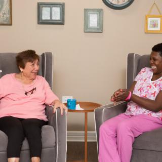 A caregiver and an elderly woman sharing a light-hearted conversation in a cozy living room setting.