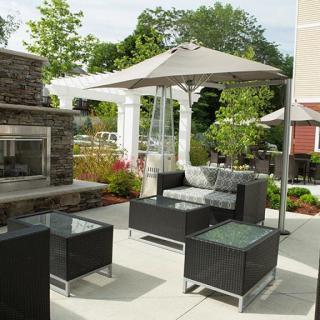 A well-designed outdoor patio with modern furniture and a fireplace, shaded by an umbrella, surrounded by lush greenery.