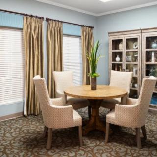 A stylish dining room with a round wooden table surrounded by four upholstered chairs. The room features elegant curtains, a decorative cabinet, and a potted plant centerpiece.