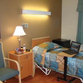 A hospital room featuring a bed, side table with a lamp, chair, and a laptop on a small table.