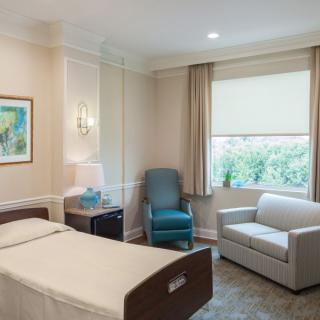 A clean and modern hospital room with a single bed, comfortable seating, and a large window.
