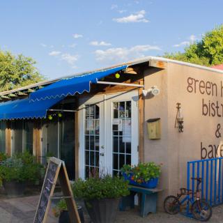 A quaint bistro and bakery with an inviting outdoor patio, featuring a blue awning and lush greenery.