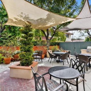 A cozy patio with tables and chairs under a shade sail, surrounded by lush greenery.