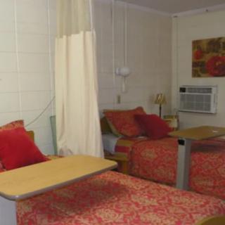 A hospital room featuring two beds with red bedding, separated by a curtain, equipped with overbed tables and an air conditioner.