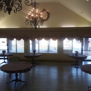 A spacious event room featuring elegant chandeliers, tables arranged for gatherings, and large windows allowing natural light.