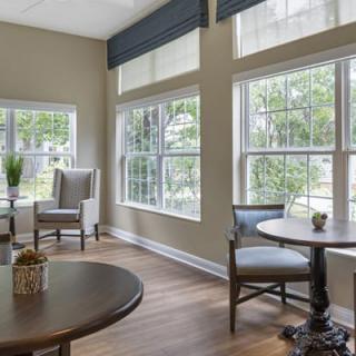 A cozy living room with large windows, allowing natural light to brighten the space, featuring comfortable chairs and round tables.