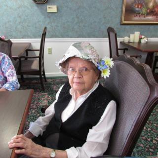 An elderly woman wearing a decorative hat sits at a table in a dining area.