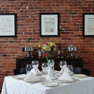 A neatly set dining table with wine glasses and white napkins against a brick wall adorned with framed art.