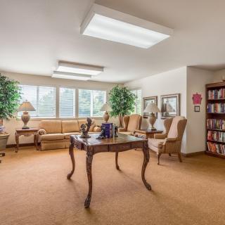 A cozy and elegantly furnished living room with a large window, featuring a sofa, chairs, bookshelf, and decorative plants.