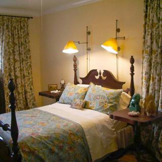 A cozy bedroom with a classic wooden bed, floral-patterned bedding and curtains, and warm lighting from wall-mounted lamps.