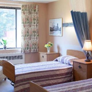 A nursing home room with two twin beds, bedside tables, a lamp, and a window with curtains.