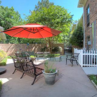 A cozy patio with outdoor seating and a large red umbrella, surrounded by greenery.