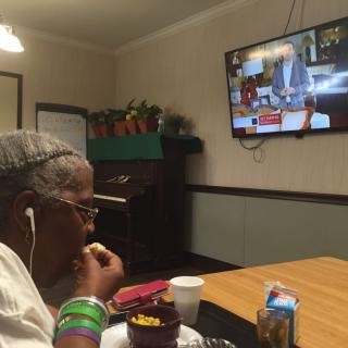 An elderly person enjoys a snack while watching television at a dining table.