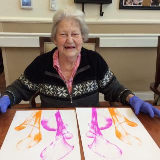 An elderly woman smiling while displaying her colorful paintings on a table.