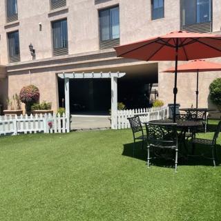 A sunny outdoor patio area with a table and chairs under an umbrella, surrounded by a green lawn and a white fence.