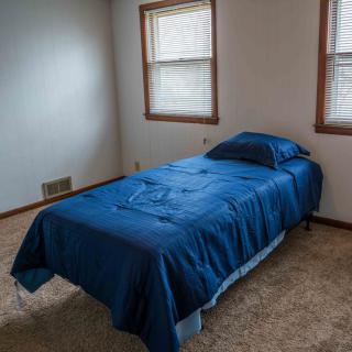 A minimalist bedroom with a single bed covered in blue bedding and two windows providing natural light.