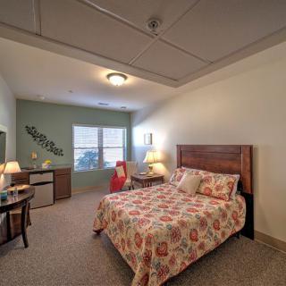 A cozy bedroom interior with a floral-patterned bedspread, a wooden headboard, ambient lighting from lamps, and a window providing natural light.