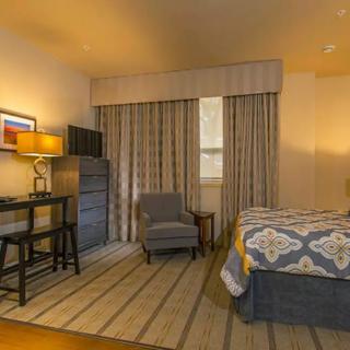 A cozy hotel room featuring a bed, armchair, desk with a lamp, and patterned curtains.