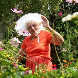 An elderly woman in a bright orange dress and sun hat enjoys her time in a vibrant flower garden.